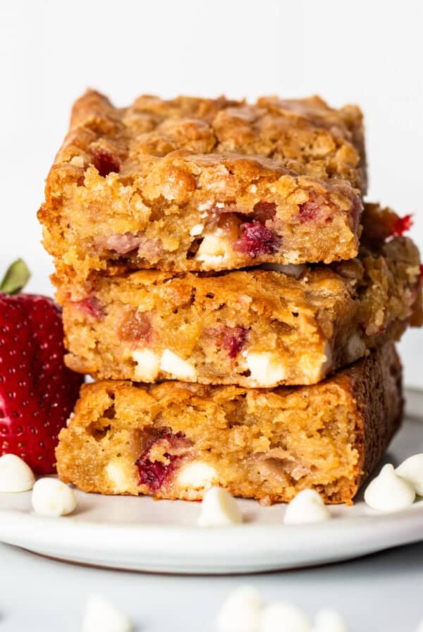 Three strawberry blondies, layered with white chocolate, are stacked on a white plate, surrounded by fresh strawberries and white chocolate chips.