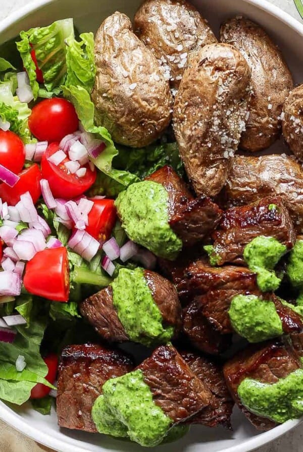 A bowl containing roasted potatoes, a fresh salad with lettuce, tomatoes, and onions, and air fryer steak bites topped with green sauce.