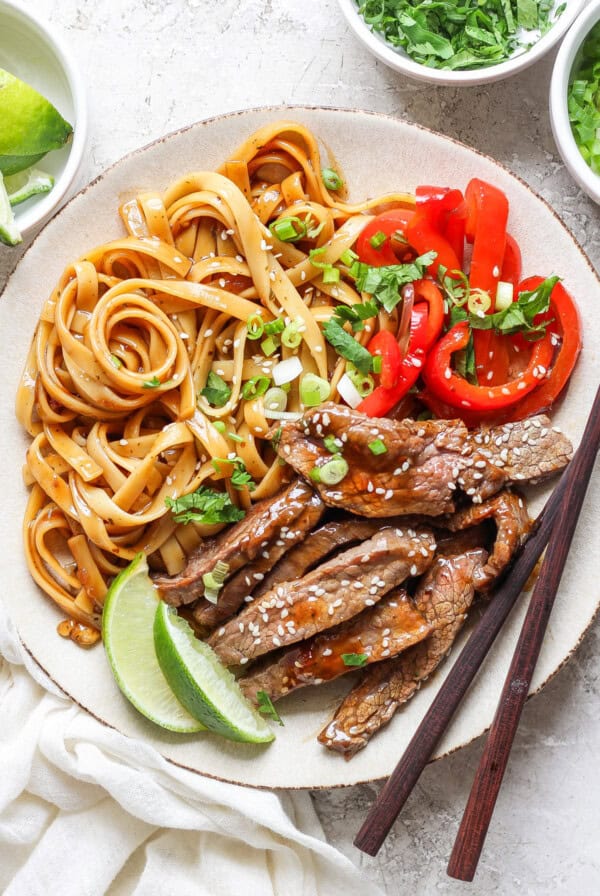 A plate with cooked noodles, sliced red bell peppers, and slices of beef topped with sesame seeds. A lime wedge is on the side, and a pair of chopsticks rests on the plate.