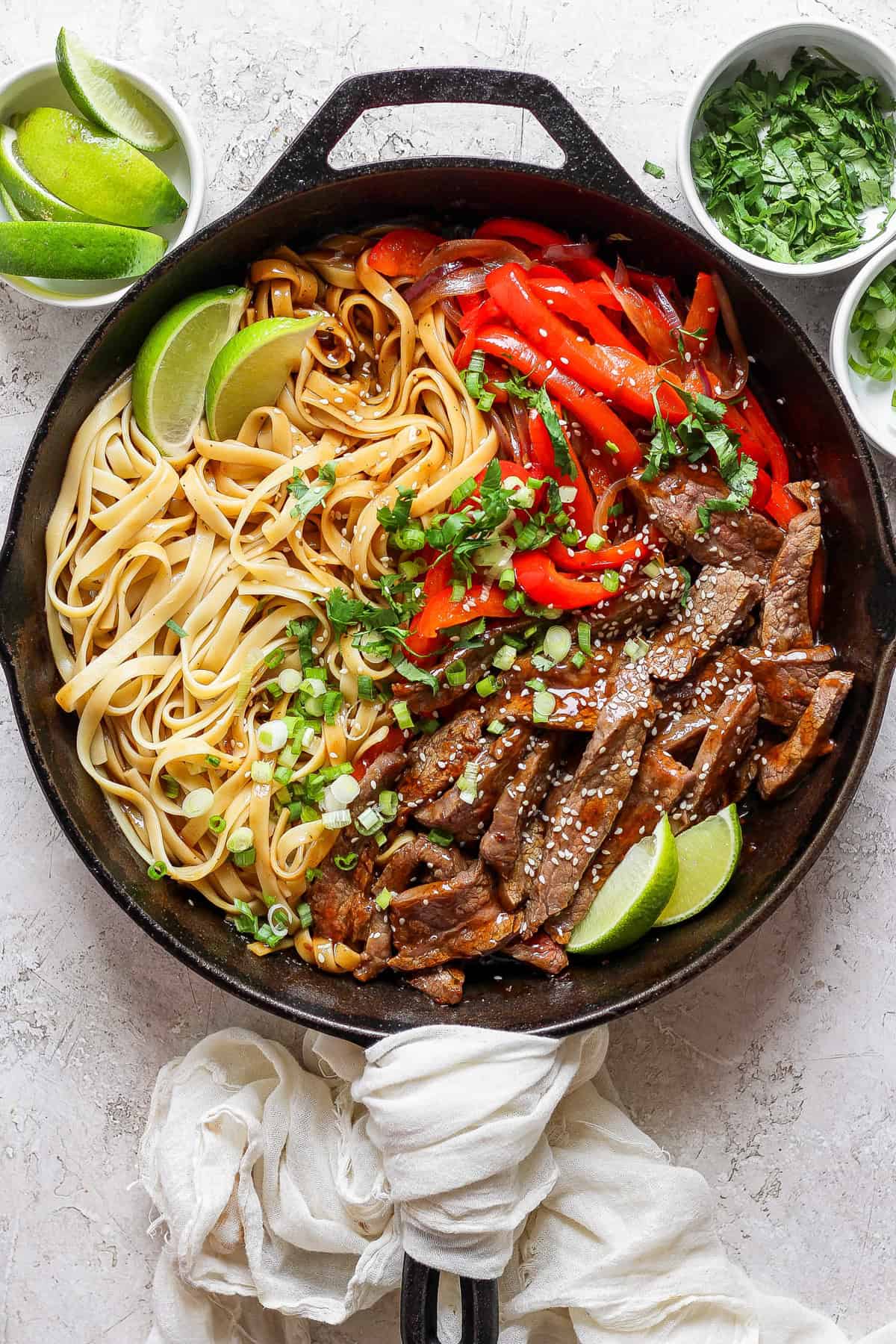 A cast iron skillet contains cooked noodles, sliced beef, red bell peppers, and lime wedges. Green herbs and lime slices are in small bowls nearby. A white cloth is draped over the skillet handle.