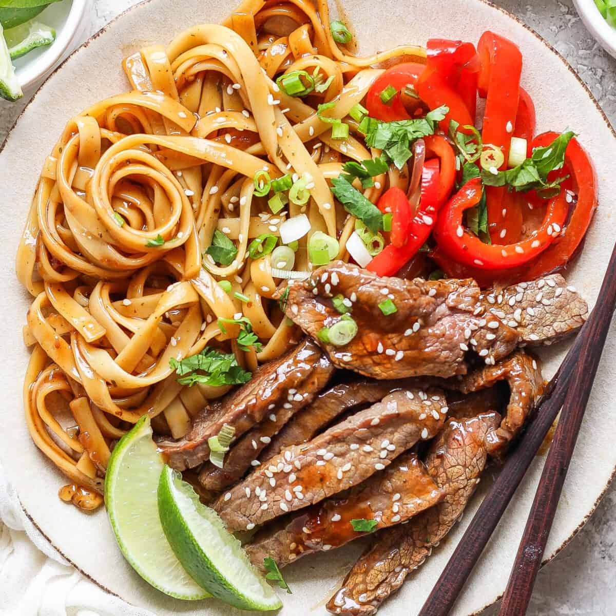 A plated dish featuring seasoned noodles, slices of red bell pepper, sesame-seeded beef strips, garnished with green onions and lime wedges, alongside a pair of chopsticks.