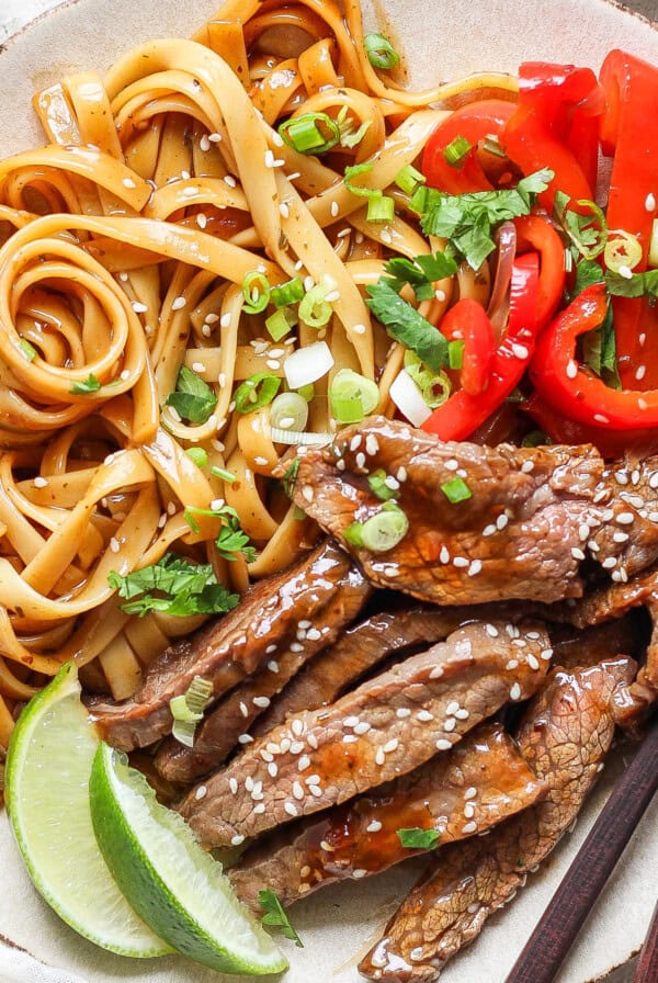 A plated dish featuring seasoned noodles, slices of red bell pepper, sesame-seeded beef strips, garnished with green onions and lime wedges, alongside a pair of chopsticks.