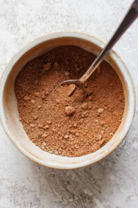 A spoon in a ceramic bowl filled with brown powder, set on a textured surface.