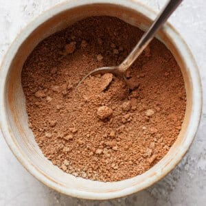 A spoon in a ceramic bowl filled with brown powder, set on a textured surface.