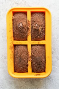 A yellow silicone baking mold with four rectangular cavities, each filled with a brown baked mixture dusted with a light coating of cocoa powder.