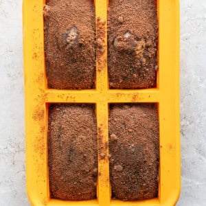 A yellow silicone baking mold with four rectangular cavities, each filled with a brown baked mixture dusted with a light coating of cocoa powder.