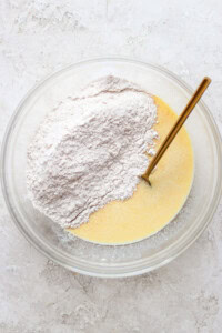 A clear bowl contains a mixture of liquid ingredients and dry flour with a gold spoon, set on a textured white surface.