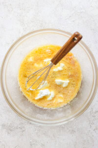 A glass bowl containing a beaten egg mixture and dollops of cream or butter, with a whisk resting inside.