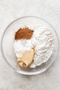 A glass bowl containing flour, brown sugar, cinnamon, baking powder, and baking soda, on a light textured surface.