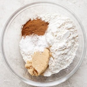 A glass bowl containing flour, brown sugar, cinnamon, baking powder, and baking soda, on a light textured surface.