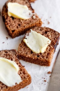 Slices of brown bread topped with thin pats of butter are placed on parchment paper next to a butter knife.