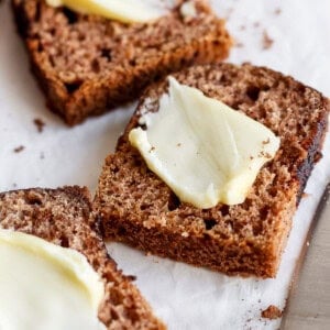 Slices of brown bread topped with thin pats of butter are placed on parchment paper next to a butter knife.