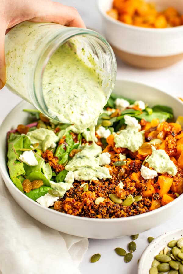 A hand pouring green herb dressing from a jar onto a bowl containing quinoa, roasted vegetables, greens, seeds, and crumbled cheese with another bowl of roasted vegetables in the background.