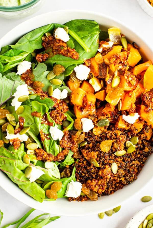 A bowl of salad featuring spinach, roasted pumpkin, quinoa, crumbled feta, pumpkin seeds, golden raisins, and a dressing drizzle.