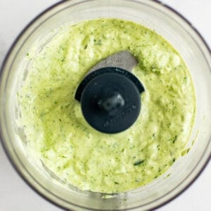 A bowl of green and white sauce sits inside a food processor with a blade in the center.