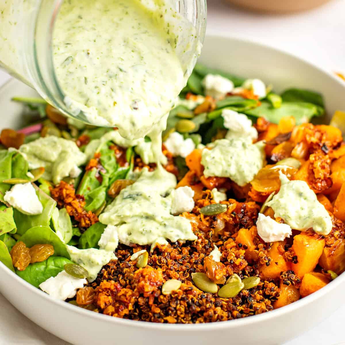 A close-up of a salad in a white bowl with quinoa, mixed greens, pumpkin seeds, raisins, and crumbled cheese, being topped with a creamy green dressing poured from a jar.