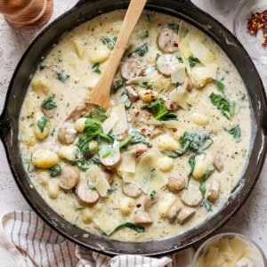 A skillet containing creamy gnocchi with sausage, spinach, and shaved cheese, with a wooden spoon and additional ingredients like spinach and cheese flakes in bowls nearby.