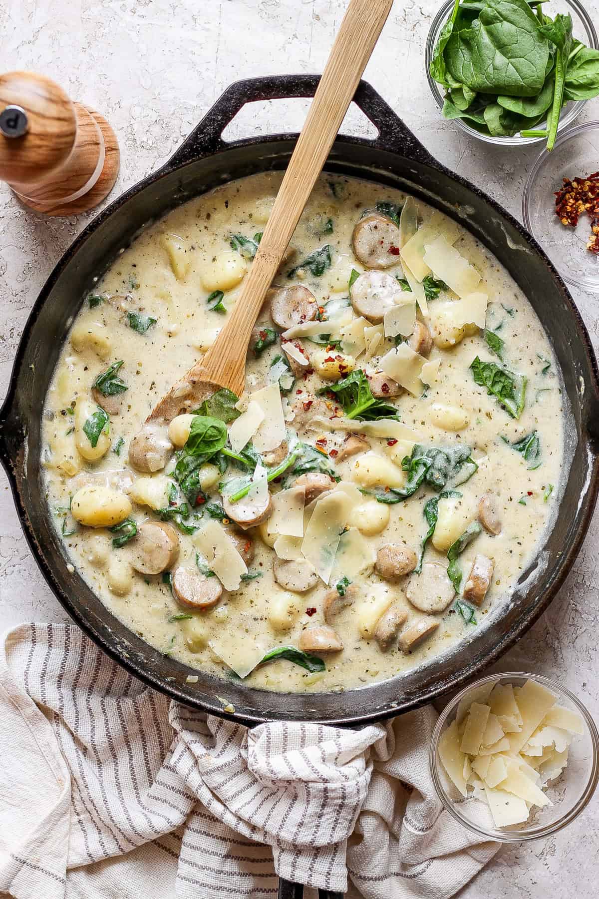 A skillet containing creamy gnocchi with sausage, spinach, and shaved cheese, with a wooden spoon and additional ingredients like spinach and cheese flakes in bowls nearby.