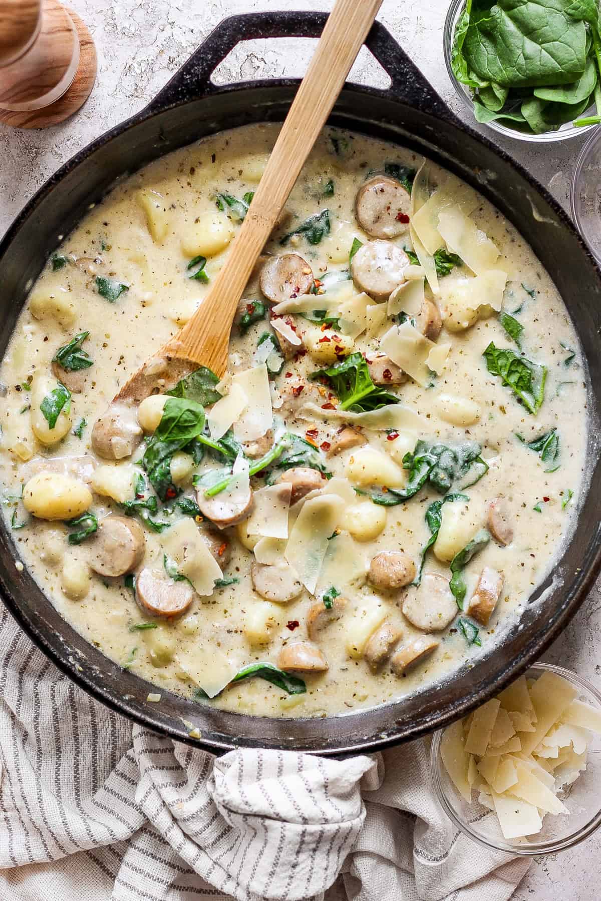 A skillet filled with creamy sauce, sliced sausage, gnocchi, spinach, and shaved cheese, with a wooden spoon resting inside. Some ingredients and a striped cloth are nearby.