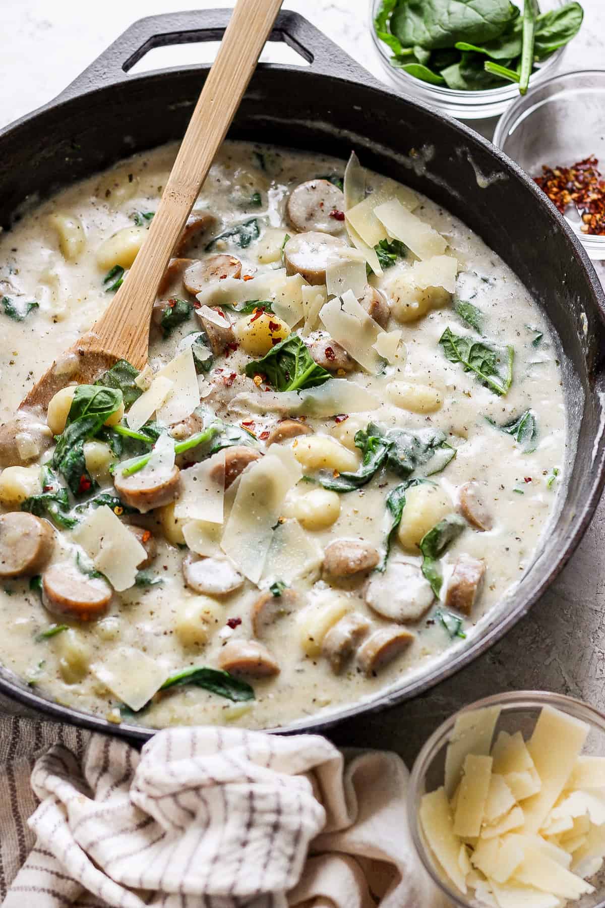 A skillet filled with creamy gnocchi, sausage slices, spinach, and topped with shaved parmesan cheese. A wooden spoon rests in the skillet, with additional ingredients visible in small bowls nearby.