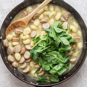 A skillet containing gnocchi, sliced sausages, and fresh spinach, with a wooden spoon resting inside.