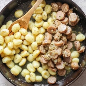 A skillet containing cooked gnocchi and sliced sausages mixed with a sauce, with a wooden spoon resting inside.