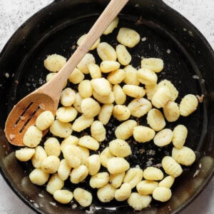 A cast iron skillet filled with cooked gnocchi, stirred with a wooden spoon, on a light gray textured surface.