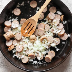 A black skillet containing sliced sausages and diced onions being stirred with a wooden spoon.