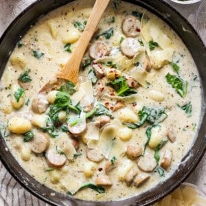 A skillet filled with creamy gnocchi, spinach, and sausage topped with shredded cheese and red pepper flakes. A wooden spoon rests inside the skillet.