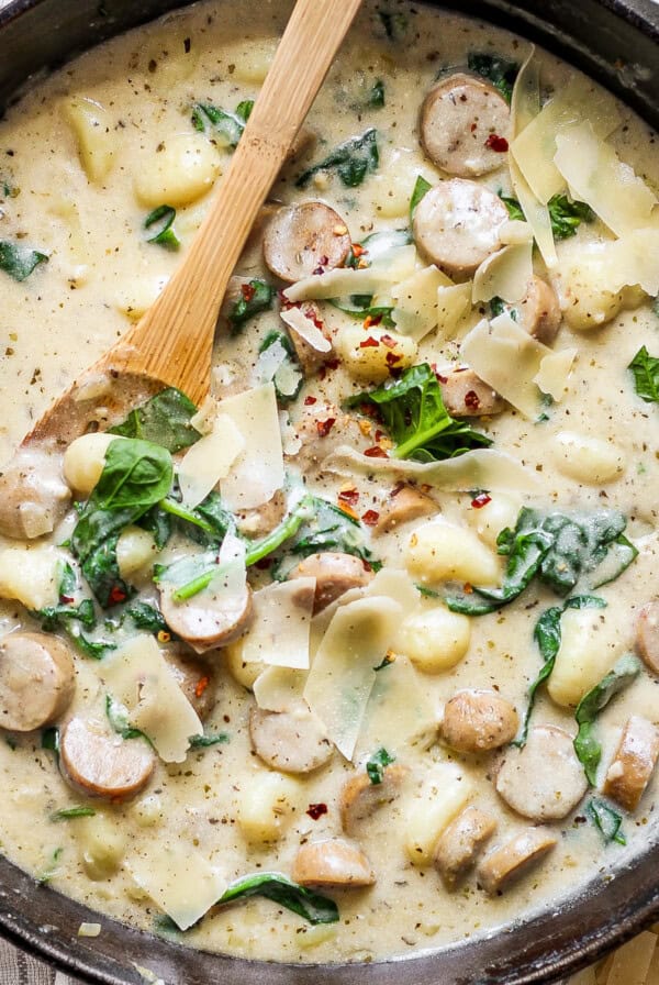 A skillet filled with creamy gnocchi, spinach, and sausage topped with shredded cheese and red pepper flakes. A wooden spoon rests inside the skillet.