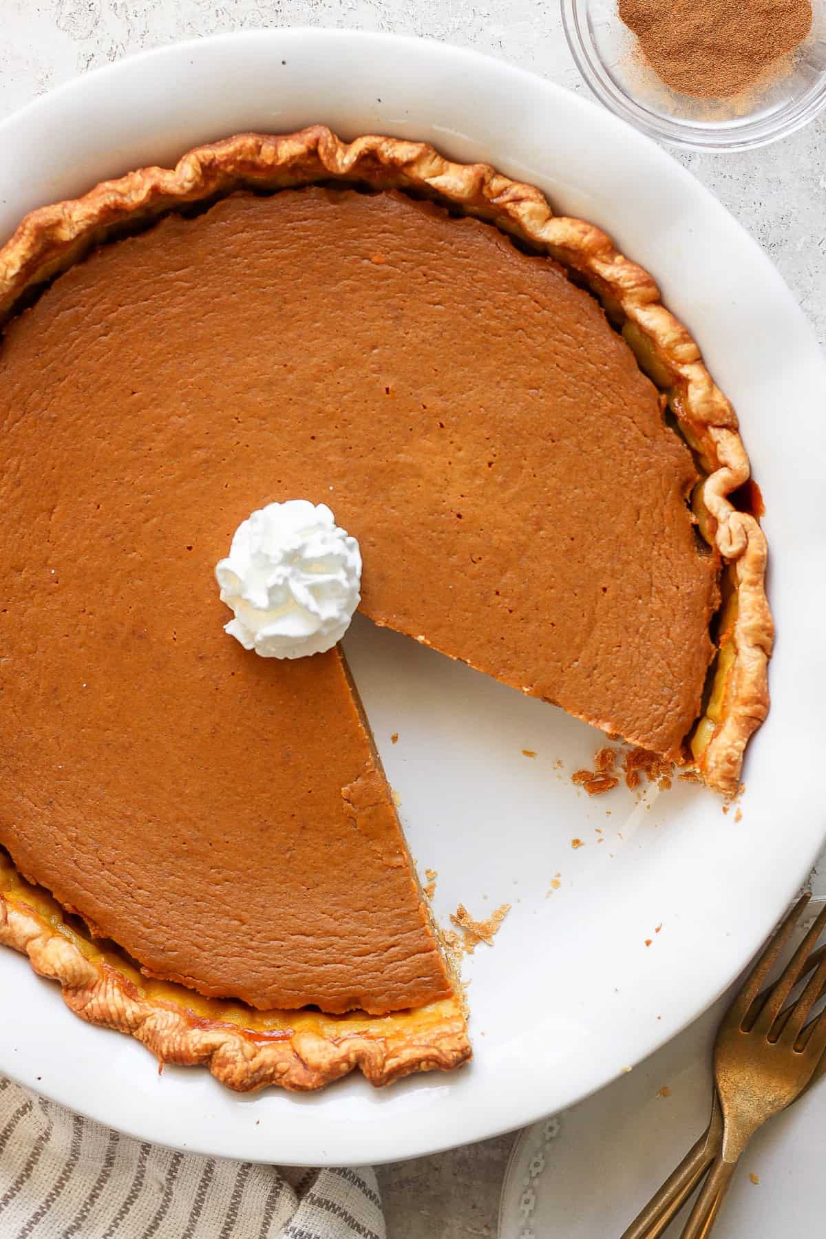 A pumpkin pie with a slice missing, topped with a dollop of whipped cream, in a round white pie dish. There is a folded towel and two gold forks next to the dish.