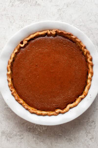 A whole pumpkin pie with a golden-brown crust in a white ceramic pie dish on a textured gray surface.