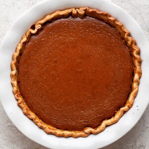 A whole pumpkin pie with a golden-brown crust in a white ceramic pie dish on a textured gray surface.