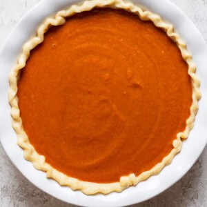 A round, unbaked pumpkin pie with a crimped edge in a white pie dish sits on a light-colored textured surface.