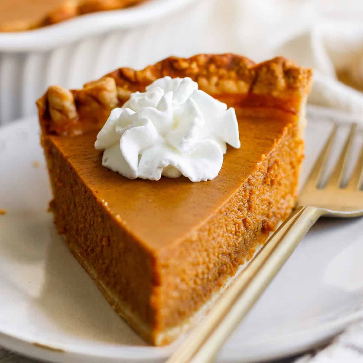 A slice of pumpkin pie topped with a dollop of whipped cream, placed on a white plate with a fork beside it.