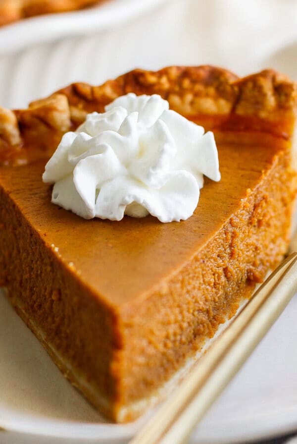 A slice of pumpkin pie topped with a dollop of whipped cream, placed on a white plate with a fork beside it.