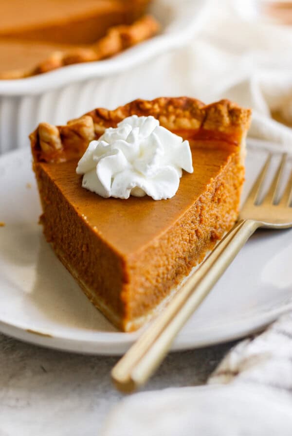 A slice of pumpkin pie topped with a dollop of whipped cream sits on a white plate with a fork placed beside it.