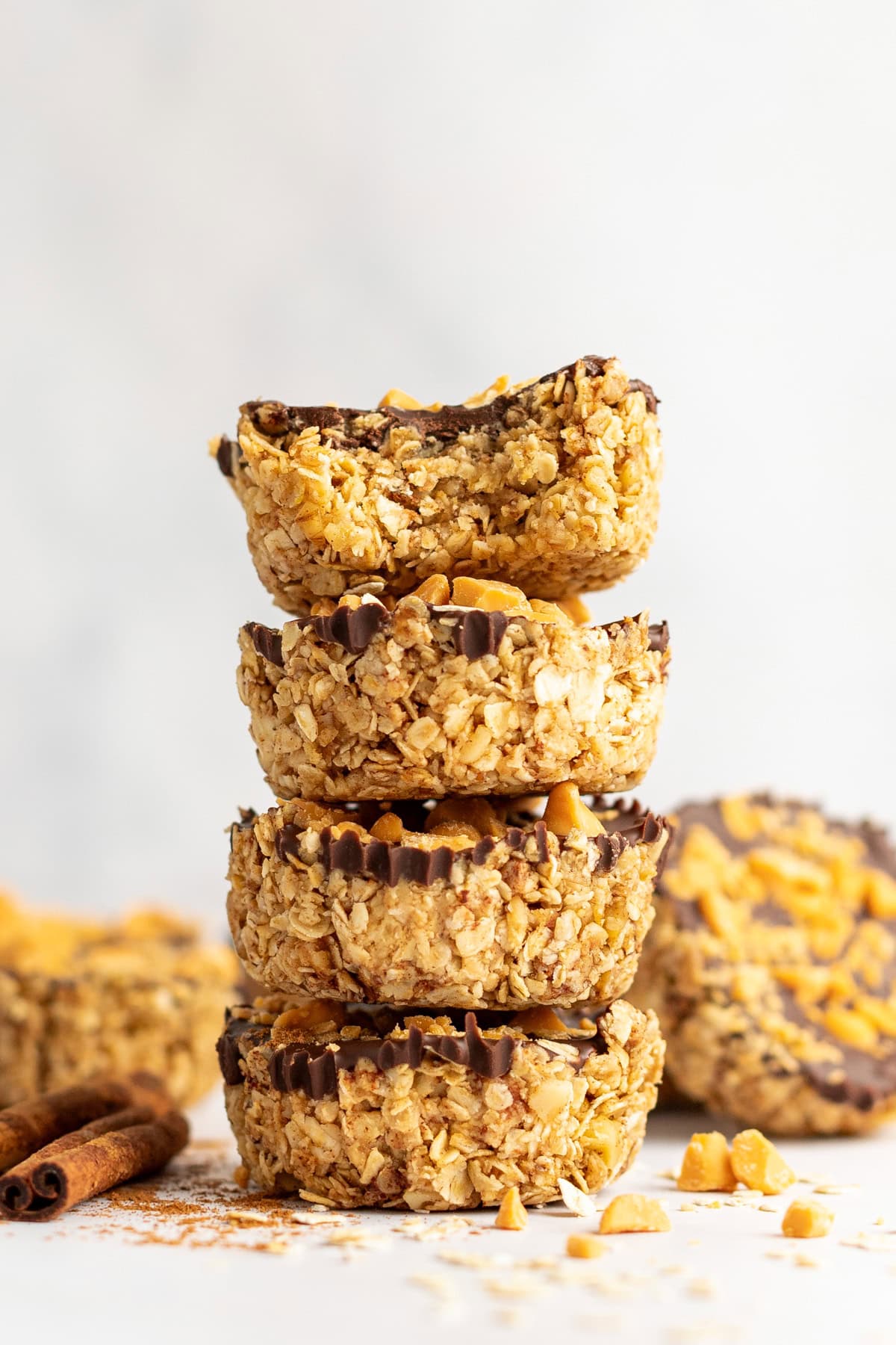 A stack of four oat and chocolate chip granola cups with a bite taken out of the top one, sitting on a light surface with scattered ingredients around.