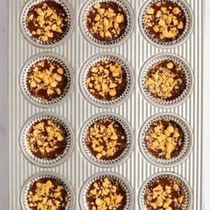 Muffin tin with twelve slots filled with chocolate batter topped with crushed nuts, lined with silver foil cupcake liners, ready for baking.