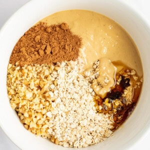 A white bowl containing peanut butter, chopped nuts, oats, cocoa powder, and honey.