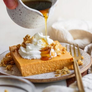 A hand pours syrup from a small jug onto a slice of cheesecake topped with whipped cream and granola. A fork and a napkin lie beside the plate.