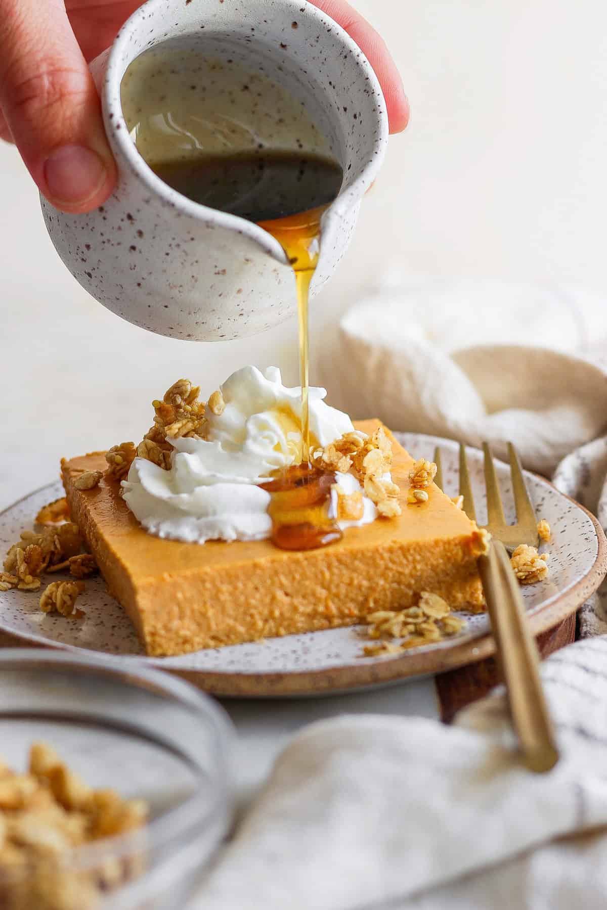 A hand pours syrup from a small jug onto a slice of cheesecake topped with whipped cream and granola. A fork and a napkin lie beside the plate.