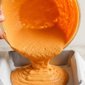 Hands pouring orange batter from a bowl into a parchment-lined baking tin.