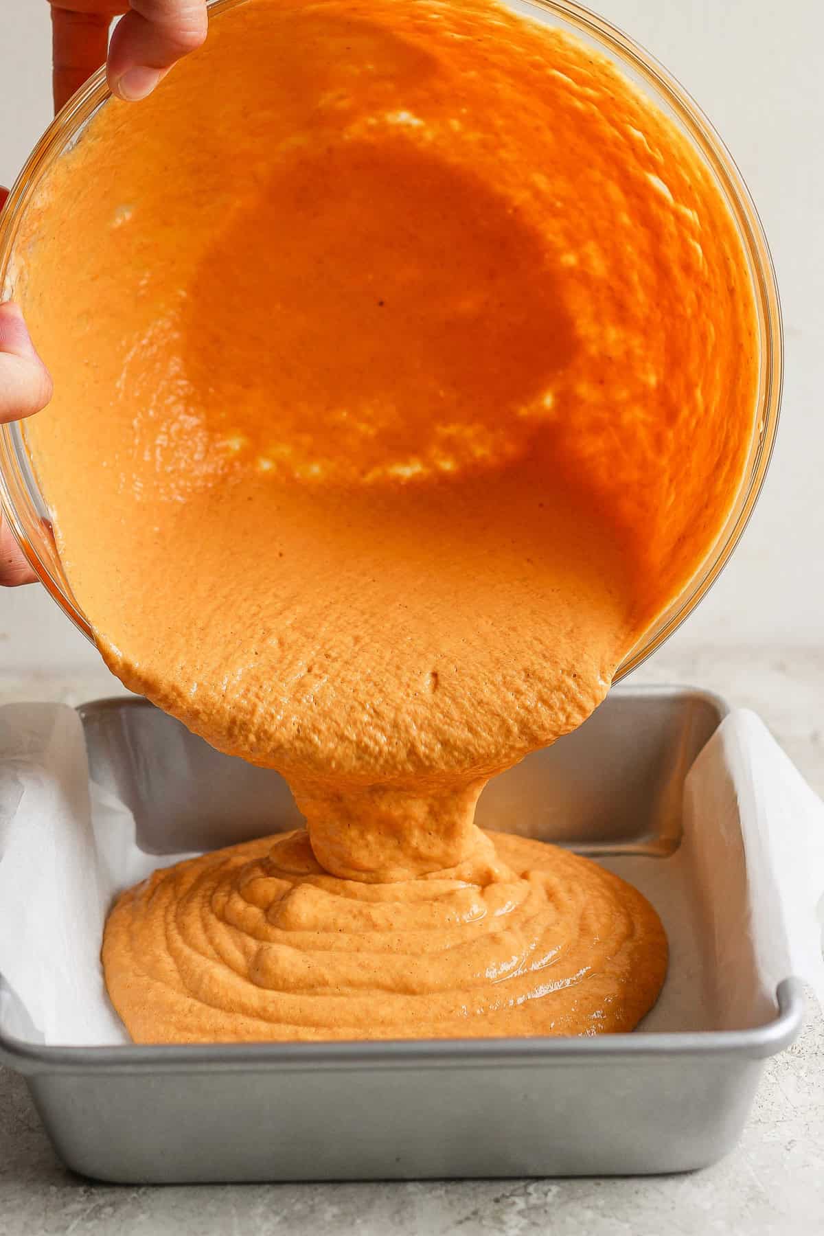 Hands pouring orange batter from a bowl into a parchment-lined baking tin.