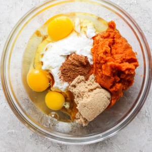 A glass mixing bowl containing raw eggs, sour cream, pumpkin puree, brown sugar, and spices, ready to be mixed.