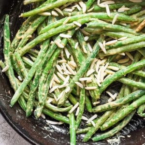 A skillet filled with green beans coated in a creamy sauce, topped with slivered almonds. A wooden spoon is visible in the top right corner.