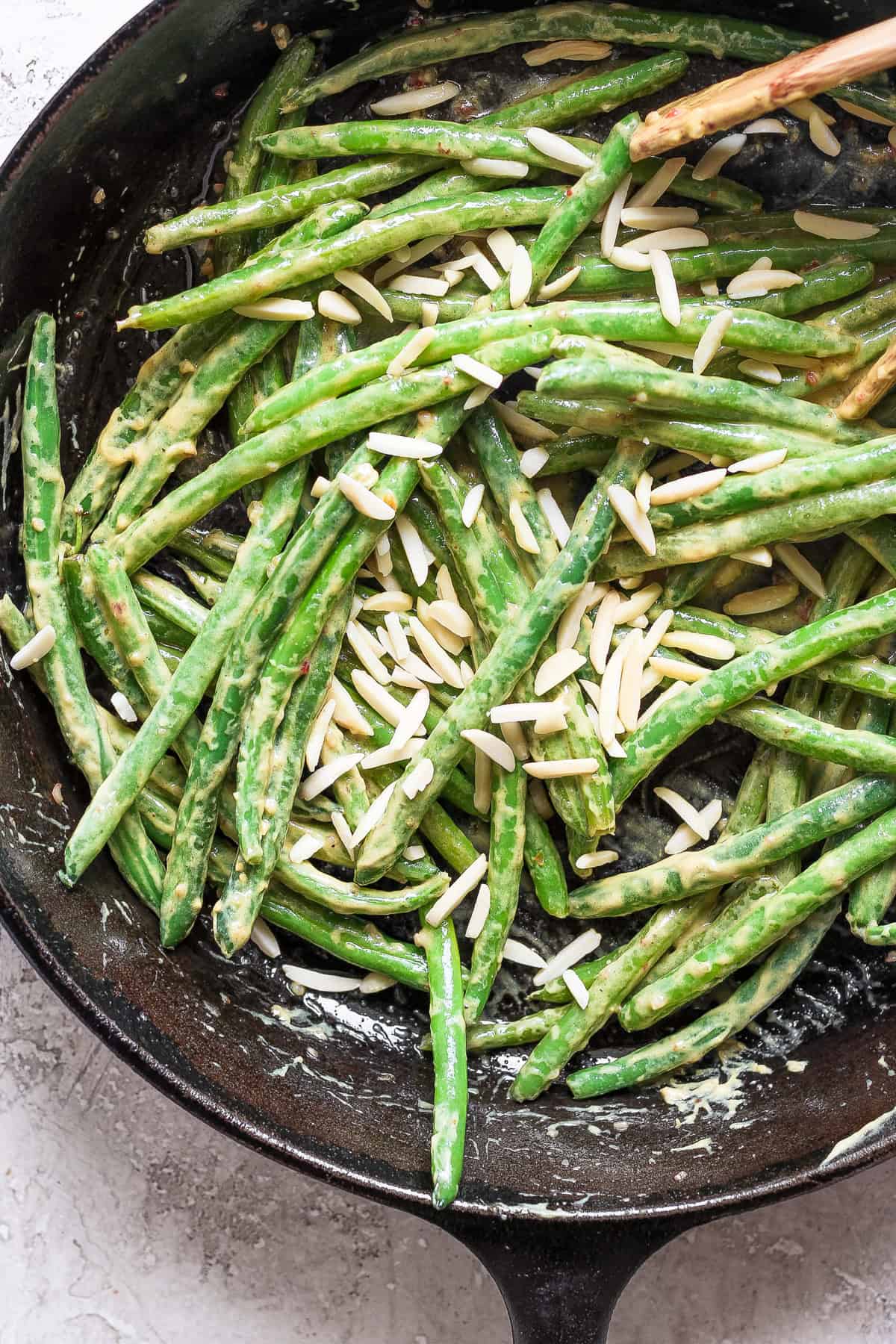 A skillet filled with green beans coated in a creamy sauce, topped with slivered almonds. A wooden spoon is visible in the top right corner.