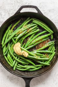 A cast iron skillet with cooked green beans, mustard, and red pepper flakes on a textured white surface.