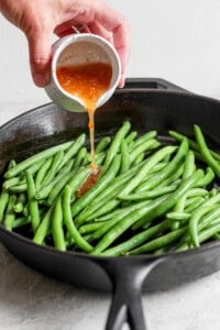 A hand pours a small container of orange sauce onto fresh green beans in a black cast iron skillet.