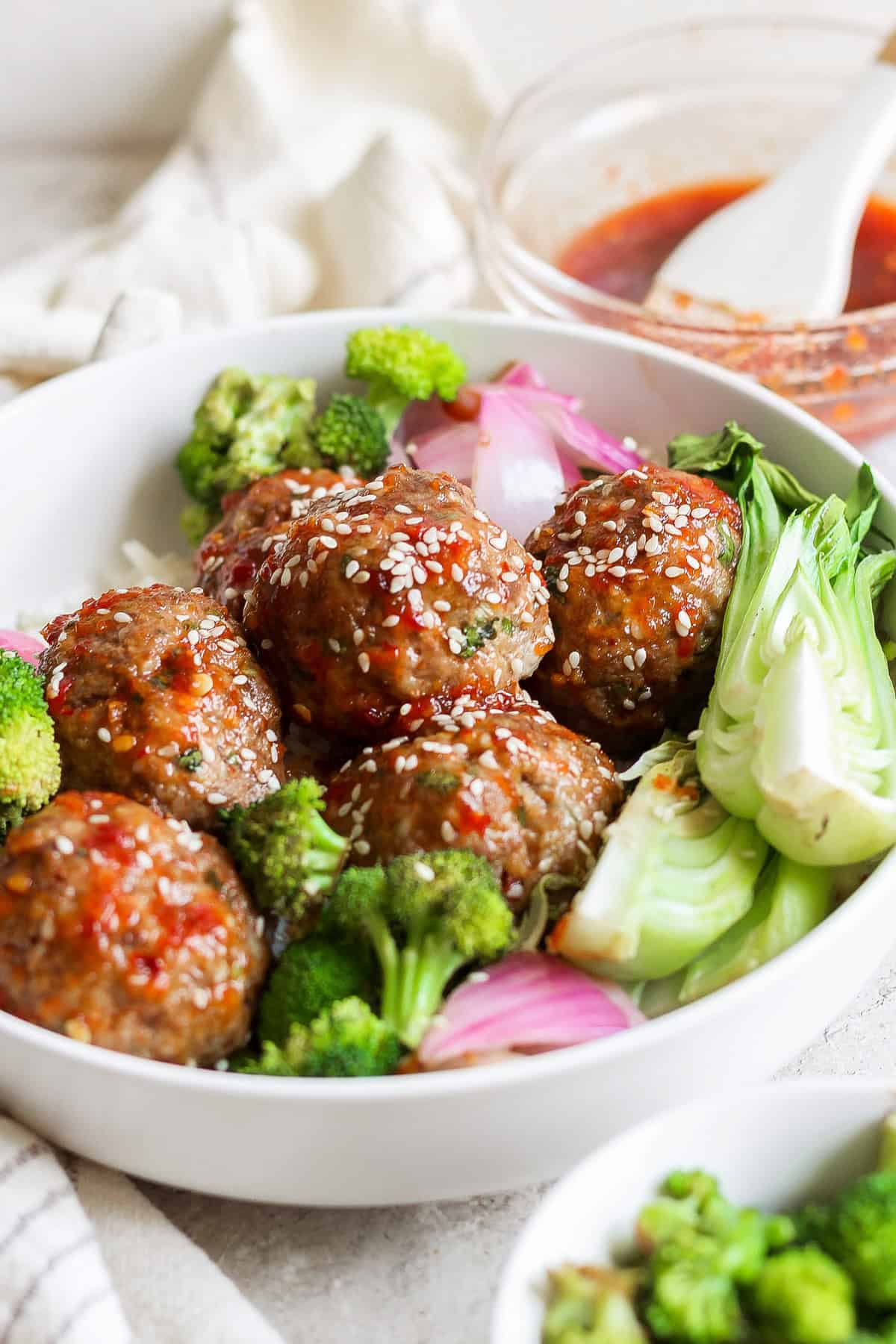 A white bowl with meatballs topped with sesame seeds and red sauce, accompanied by broccoli, bok choy, and red onions. A small dish with extra sauce and a brush is in the background.